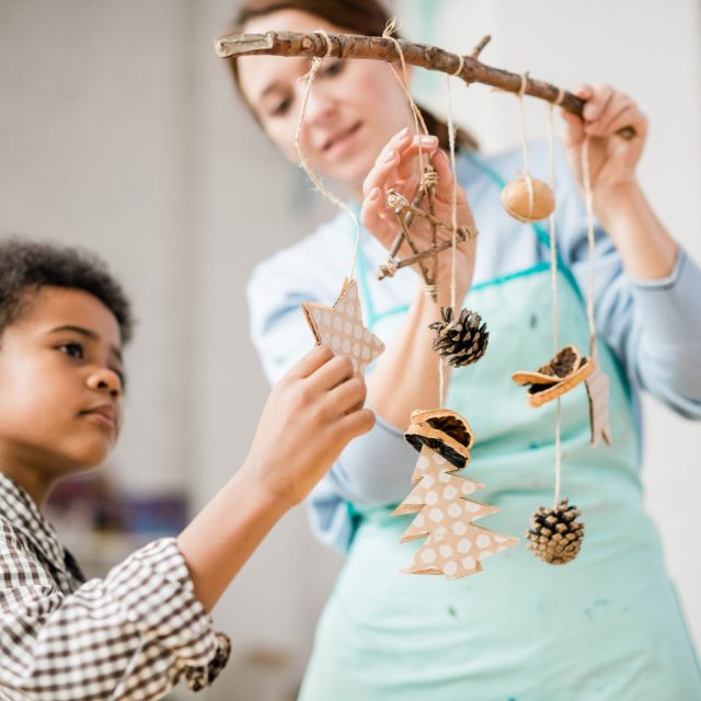 Cute boy holding paper star decoration hanging on stick among pinecones and other handmade Christmas toys